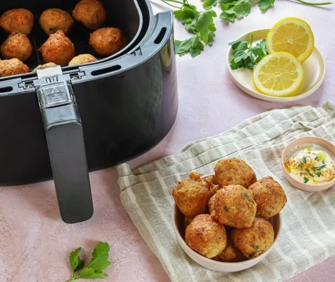 ATELIER ACRAS DE MORUE À VOLONTÉ servi dans une friteuse, avec un bol sur un tissu rayé, une sauce à tremper, et une assiette de tranches de citron et de persil sur une surface rose.