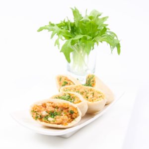 Quatre moitiés de MINI PITA POULET (Barquette de 20 pièces) remplies de poulet, de légumes et d'herbes sont disposées sur une assiette blanche. À l'arrière-plan, un verre contenant de la roquette verte fraîche est posé sur une surface blanche.