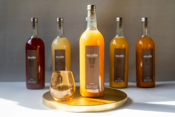 Une bouteille de NECTAR DE FRAISES ALAIN MILLIAT 1L et un verre sont posés sur un plateau rond et doré au premier plan, tandis que quatre autres nectars de fruits sont alignés derrière. La lumière du soleil met en valeur la bouteille centrale de nectar de fraise et le verre.