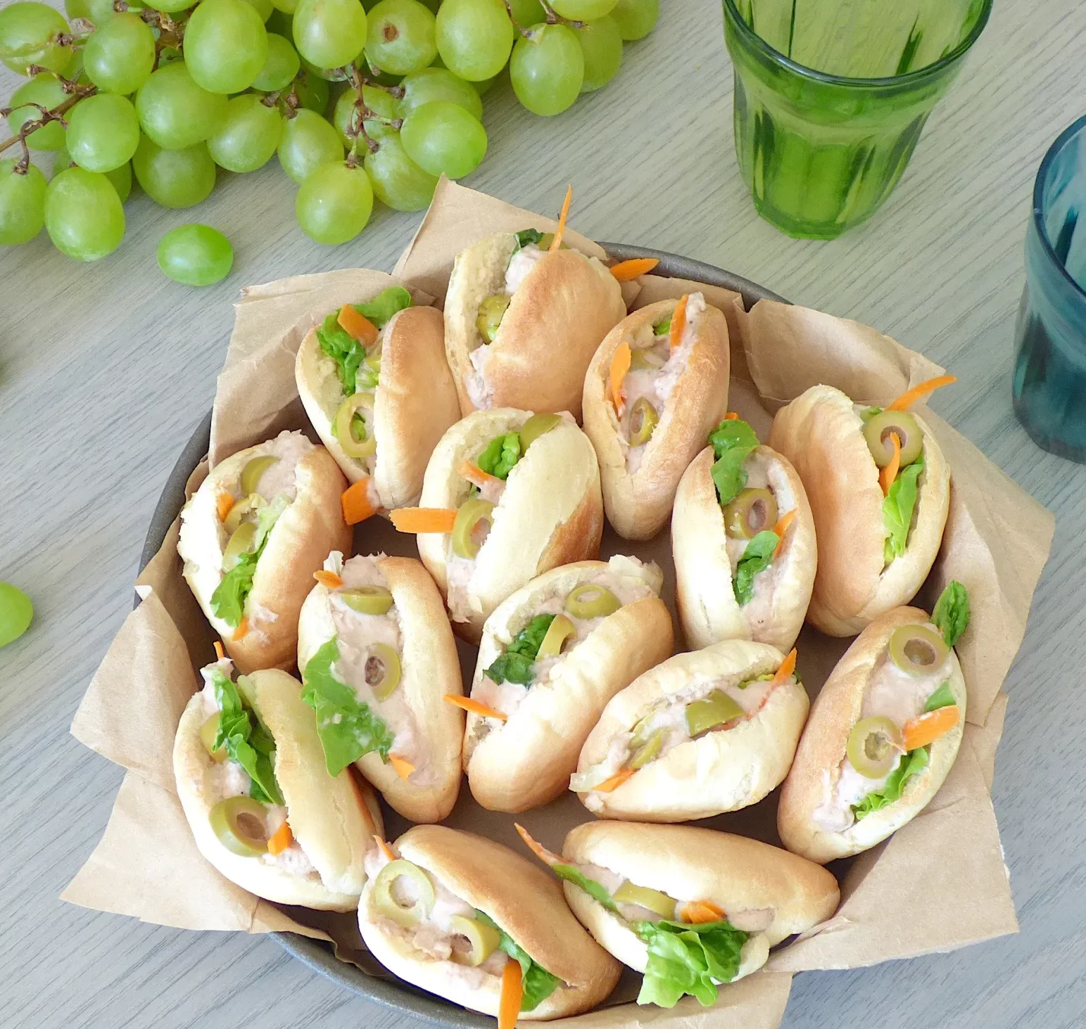 Un panier de COCKTAIL STREET FOOD (17 pièces salées et sucrées) est posé sur une table, comprenant des petits pains assortis avec de la laitue, des olives, des carottes et une garniture crémeuse, accompagnés de raisins verts et de verres à boire verts.