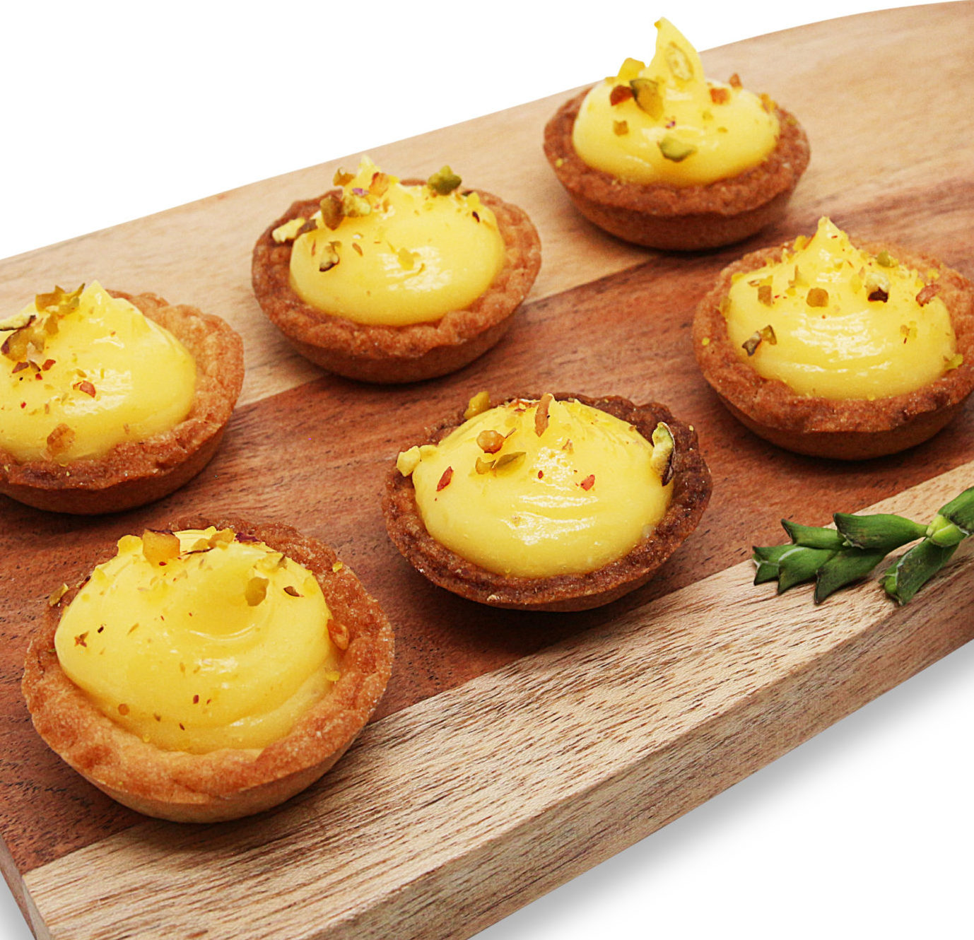 Six tartelettes sucrées de l'assortiment COCKTAIL STREET FOOD (17 pièces salées et sucrées) sont garnies d'une crème jaune et d'une garniture à la pistache. Elles sont disposées sur une planche en bois avec une petite plante verte en guise de décoration.