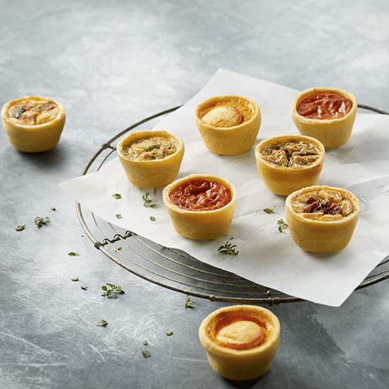 Dix-huit PETIT COCKTAIL MAGNOLIA assortis, salés et sucrés, sont présentés sur du parchemin sur une grille, garnis d'herbes sur un fond texturé gris clair.