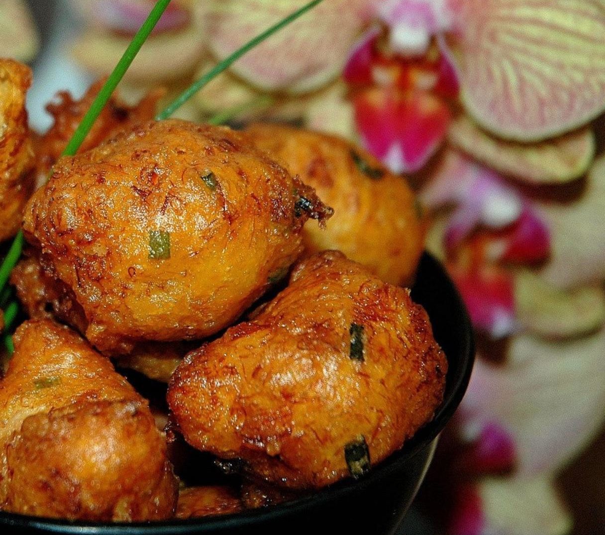 Des beignets d'ACRAS DE MORUE dorés (plateau de 30 pièces, 12 g chacune) aux herbes vertes sont disposés dans un bol devant des orchidées roses et blanches floues, et semblent croustillants et fraîchement cuits.