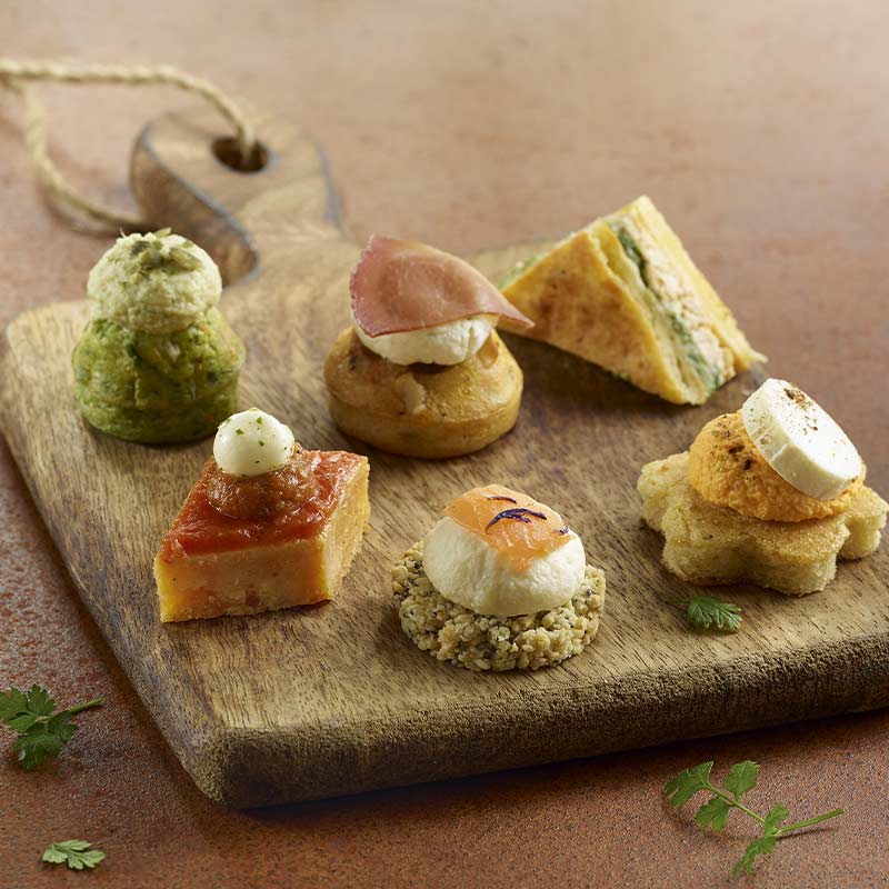 Une planche en bois présente six canapés assortis de PETIT COCKTAIL MAGNOLIA (18 pièces salées et sucrées), chacun avec des garnitures et des bases uniques, disposés avec art sur un fond brun.