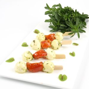 Une assiette rectangulaire blanche présente BROCHETTE ITALIENNE (Plateau de 20 pièces) : brochettes avec boules de mozzarella, tomates cerises et herbes, garnies d'herbes vertes fraîches et de petites feuilles.