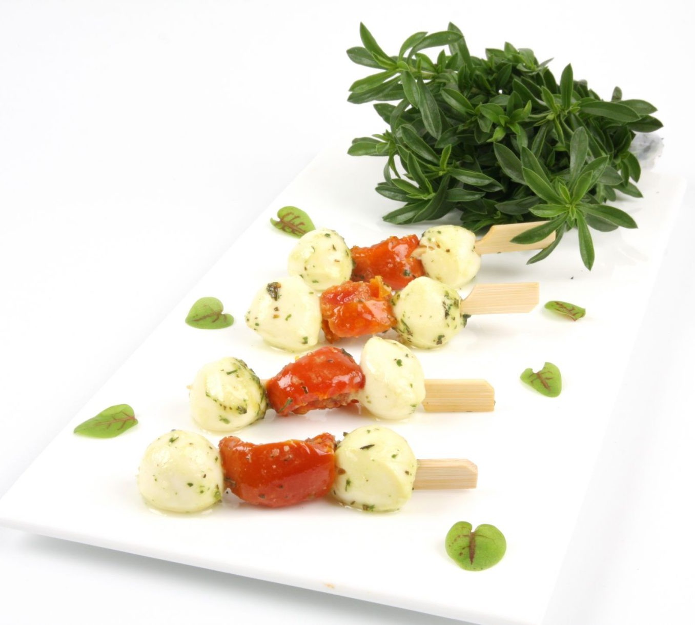 Une assiette rectangulaire blanche présente BROCHETTE ITALIENNE (Plateau de 20 pièces) : brochettes avec boules de mozzarella, tomates cerises et herbes, garnies d'herbes vertes fraîches et de petites feuilles.