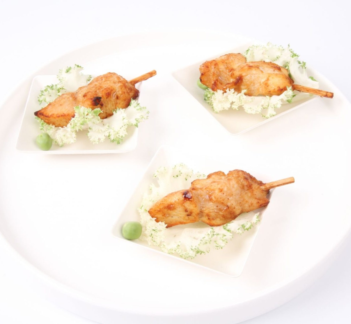 Trois brochettes de poulet BROCHETTE POULET PIC & CHIC (d'un paquet de 20 pièces) sont posées sur des lits de laitue frisée, chacune avec un pois vert, servies dans des petites assiettes disposées sur un plateau rond blanc sur fond blanc.