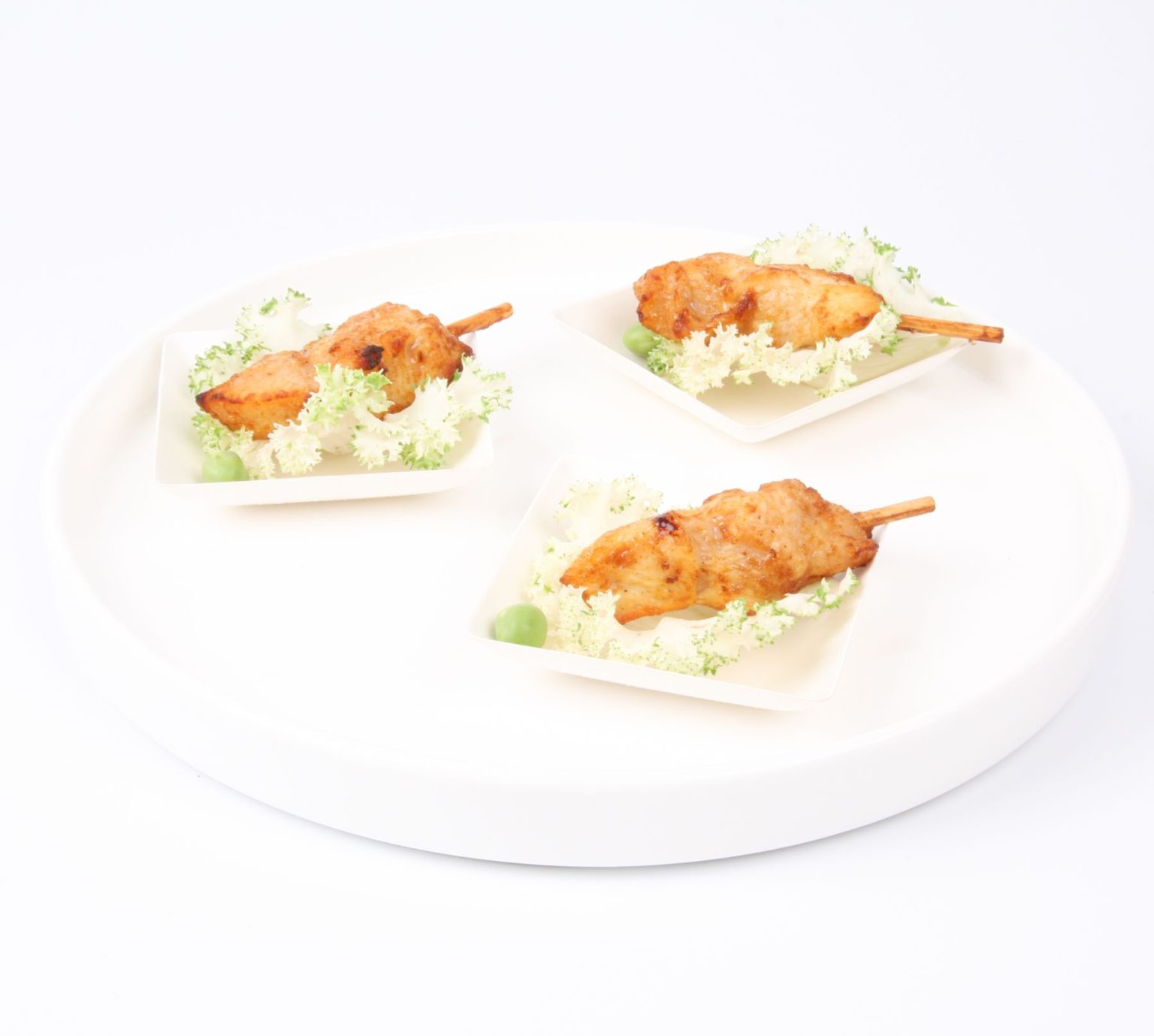 Trois brochettes de BROCHETTE POULET PIC & CHIC (Barquette de 20 pièces) sont servies dans de petites assiettes sur des feuilles vertes et garnies de petits pois, présentées sur un plateau rond blanc sur un fond blanc minimaliste.