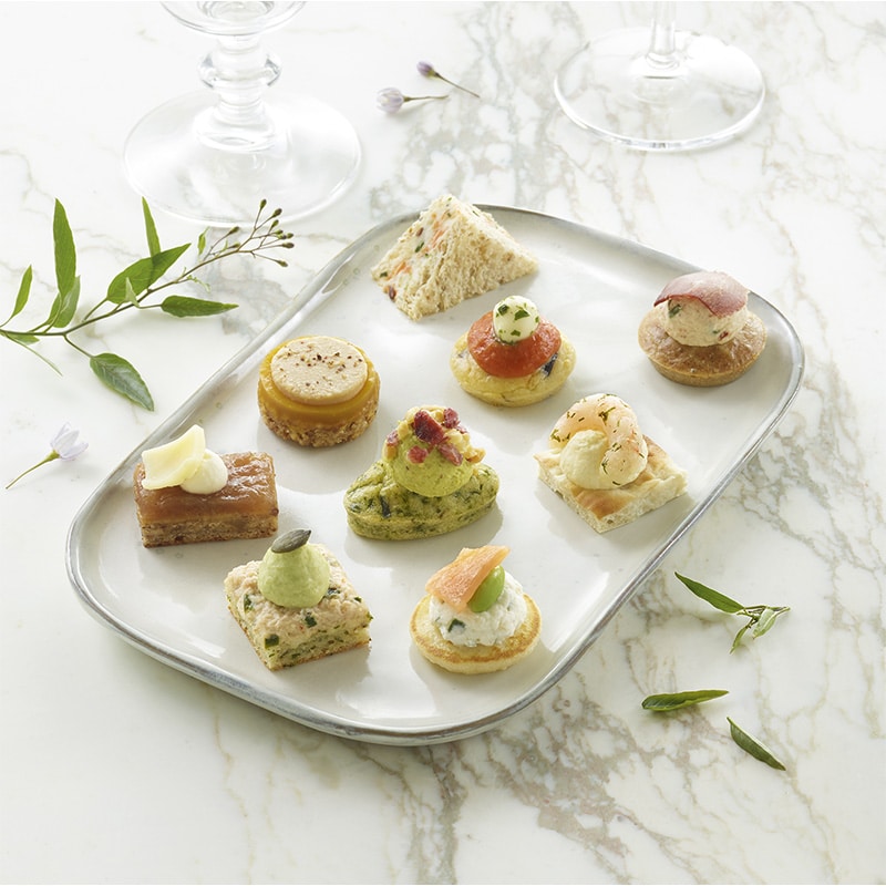 Le PETIT COCKTAIL MAGNOLIA comprend 18 bouchées salées et sucrées disposées sur un plateau, chacune garnie de façon unique, présentées sur une surface en marbre avec de la verdure et de la verrerie à proximité.