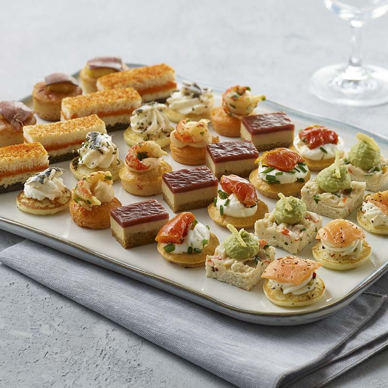 Le PETIT COCKTAIL MAGNOLIA propose 18 mini-bouchées salées et sucrées assorties, y compris des sandwichs, des tartes et des pâtisseries, joliment disposées sur un plateau blanc au-dessus d'une serviette grise pliée.