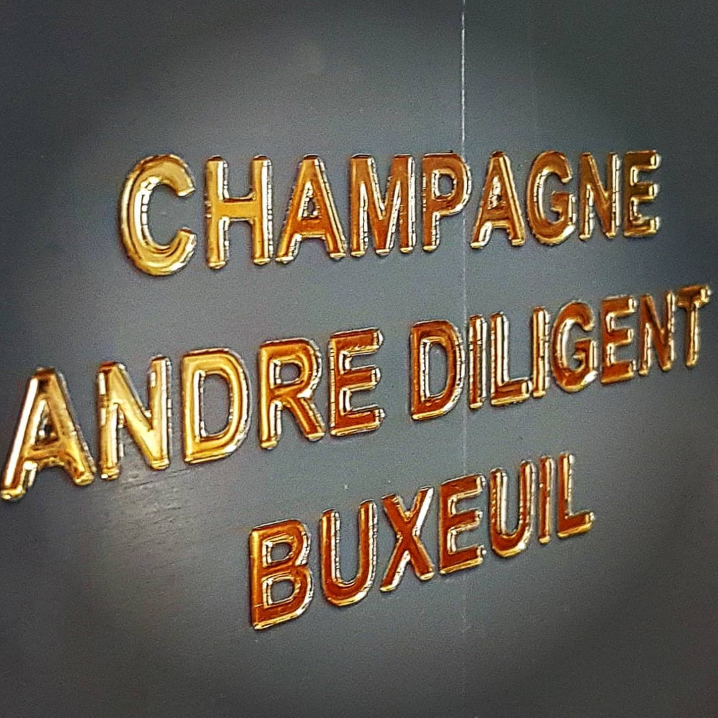 Sur un mur gris, des lettres dorées en 3D indiquent CHAMPAGNE ANDRÉ DILIGENT (Brut Blanc 75cl).