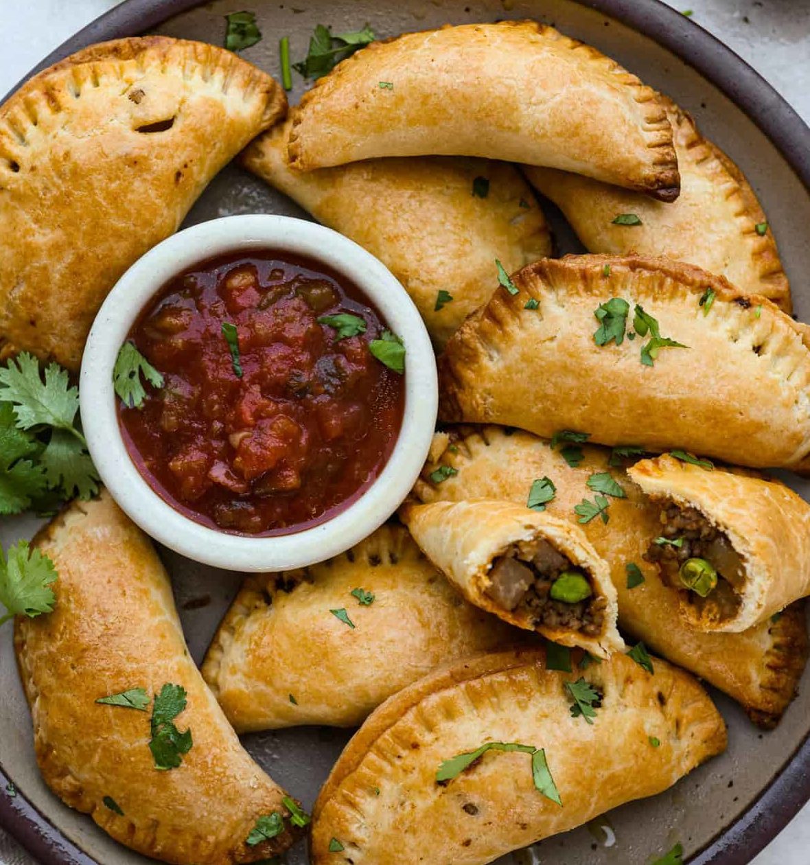 Un plateau de COCKTAIL STREET FOOD (17 pièces salées et sucrées), comprenant des empanadas dorés garnis de coriandre autour d'un bol de salsa épaisse ; un empanada est ouvert pour révéler sa garniture savoureuse.