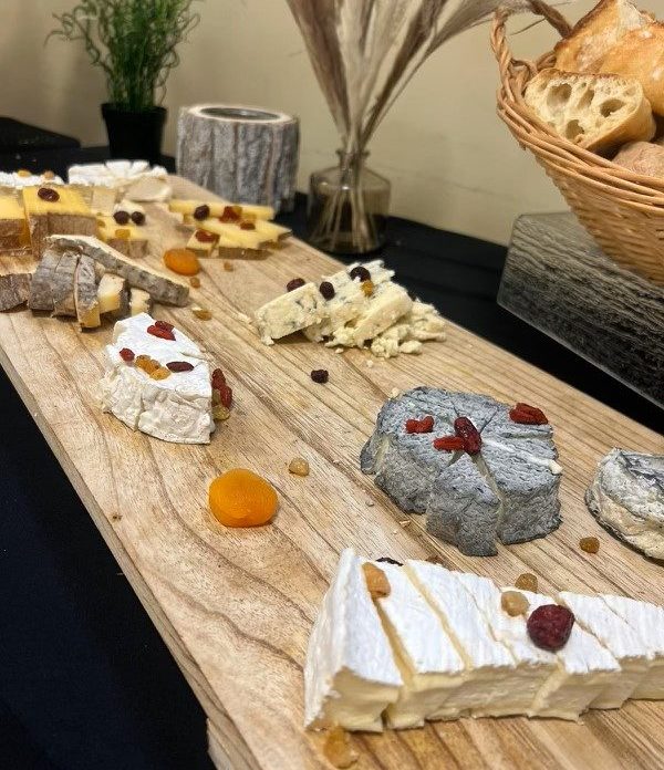 Le PLATEAU DE FROMAGE présente un assortiment de fromages garnis de canneberges et d'abricots séchés, servis sur une planche en bois avec des noix et des fruits secs. Une corbeille de pain et des plantes décoratives se trouvent à l'arrière-plan.