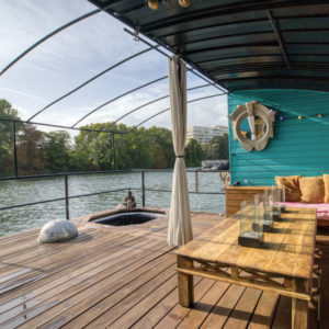 LE LOFT FLOTTANT DE NICOLAS (92200) dispose d'une terrasse en bois avec une longue table, des banquettes rembourrées, des guirlandes lumineuses colorées et un miroir rond, surplombant une rivière calme bordée d'arbres et de bâtiments urbains.