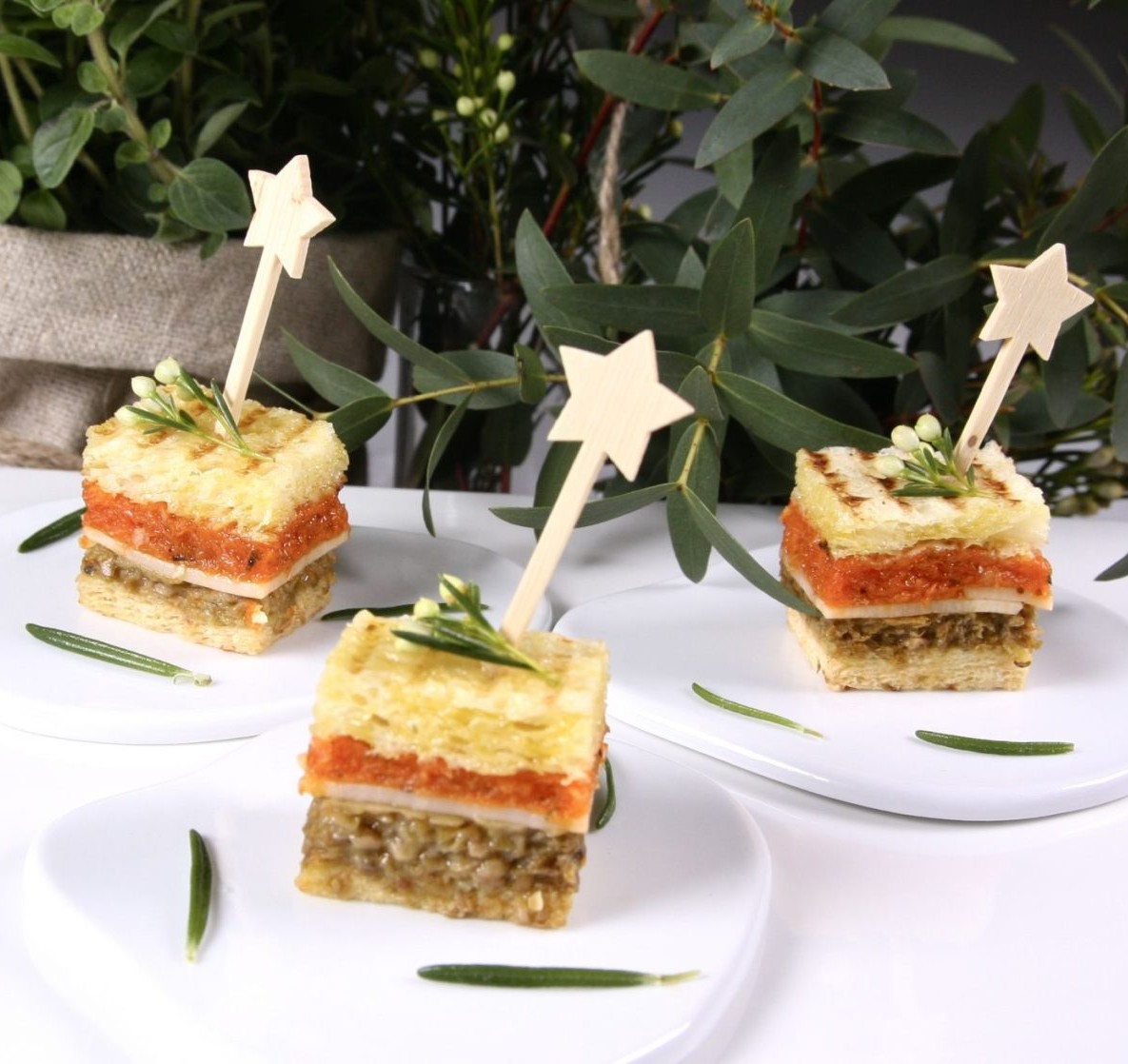 Le GRAND COCKTAIL MAGNOLIA ("BEST" 22 pièces salées et sucrées) présente trois amuse-gueules carrés superposés, chacun sur une assiette blanche avec une garniture de romarin et des légumes verts en arrière-plan, embrochés à l'aide de bâtonnets en forme d'étoiles.