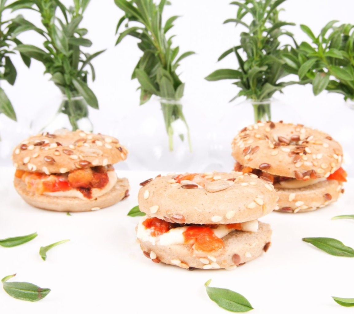 Un plateau de MINI BAGEL LEGUMES DU SOLEIL (Plateau de 20 pièces), garni de fromage et de tomates, est présenté sur une surface blanche avec des feuilles vertes, tandis que des bocaux en verre à l'arrière-plan contiennent des brins d'herbes fraîches.