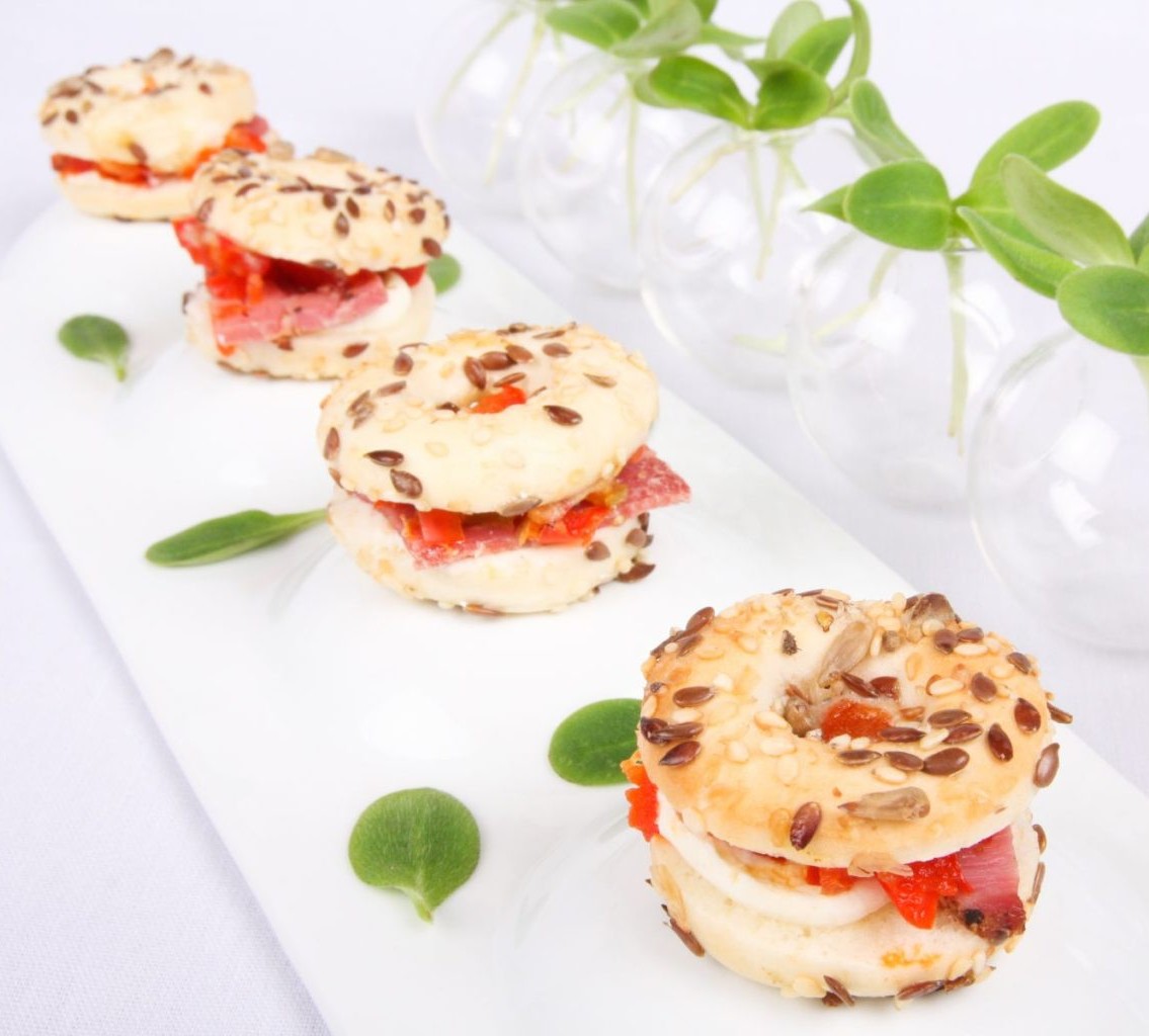 Les MINI BAGELS PASTRAMI (Plateau de 20 pièces) sont disposés en rang sur une assiette blanche, garnis de pastrami, de fromage et de poivrons rouges, décorés de feuilles vertes et de petits vases avec des tiges vertes en arrière-plan.