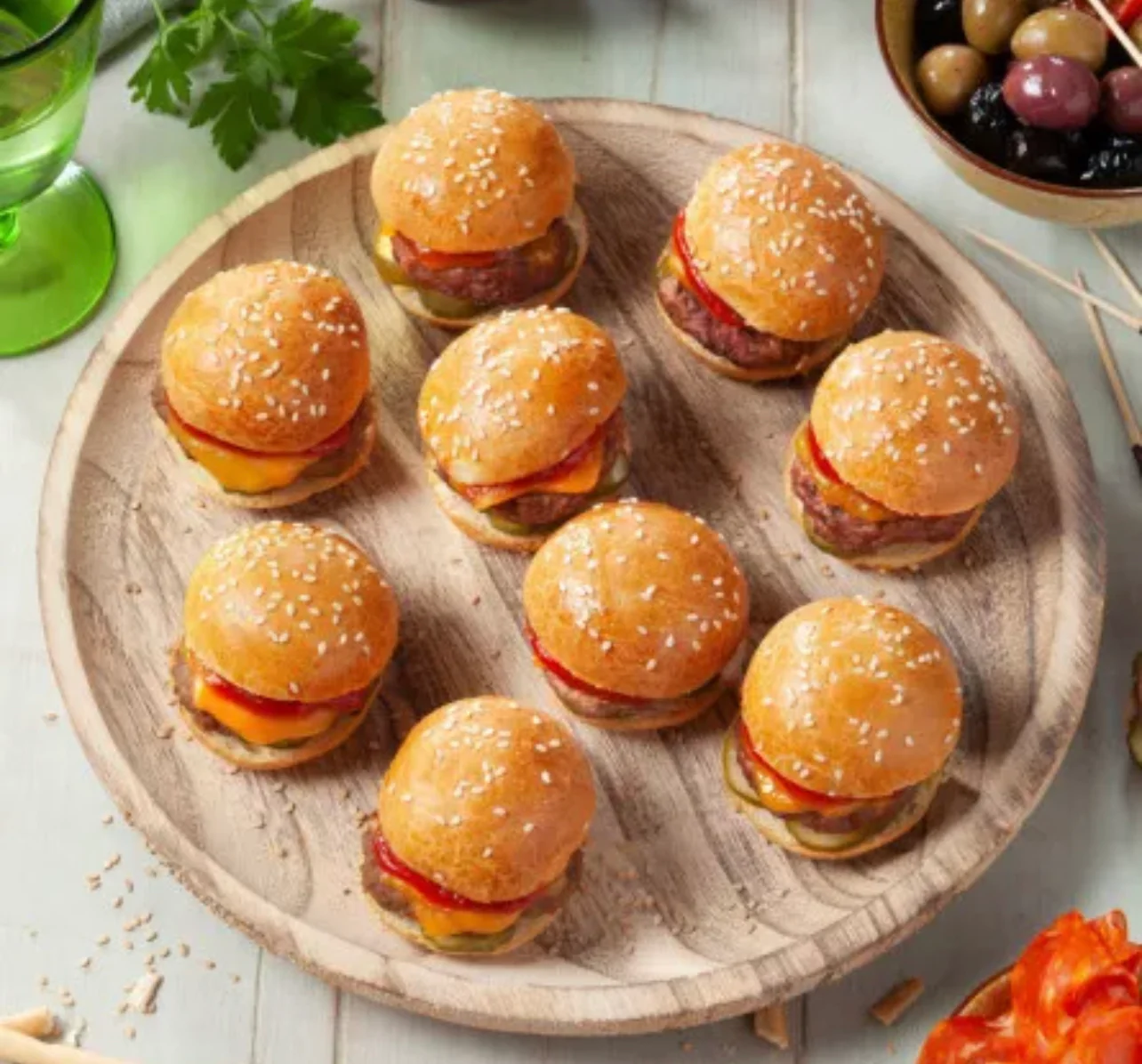 Une assiette en bois clair présente le COCKTAIL STREET FOOD (17 pièces salées et sucrées), composé de neuf mini cheeseburgers avec des petits pains aux graines de sésame, du fromage et des tranches de tomate. Des olives et du persil apparaissent en arrière-plan sur une table lumineuse.