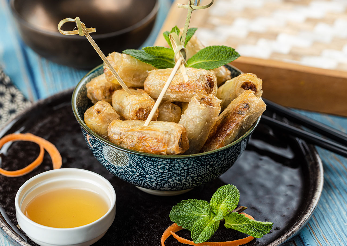 COCKTAIL STREET FOOD (17 pièces salées et sucrées) présenté avec des rouleaux de printemps croustillants, garnis de menthe et servis avec une sauce dip et des décorations de carotte sur une assiette foncée, accompagnés de baguettes.