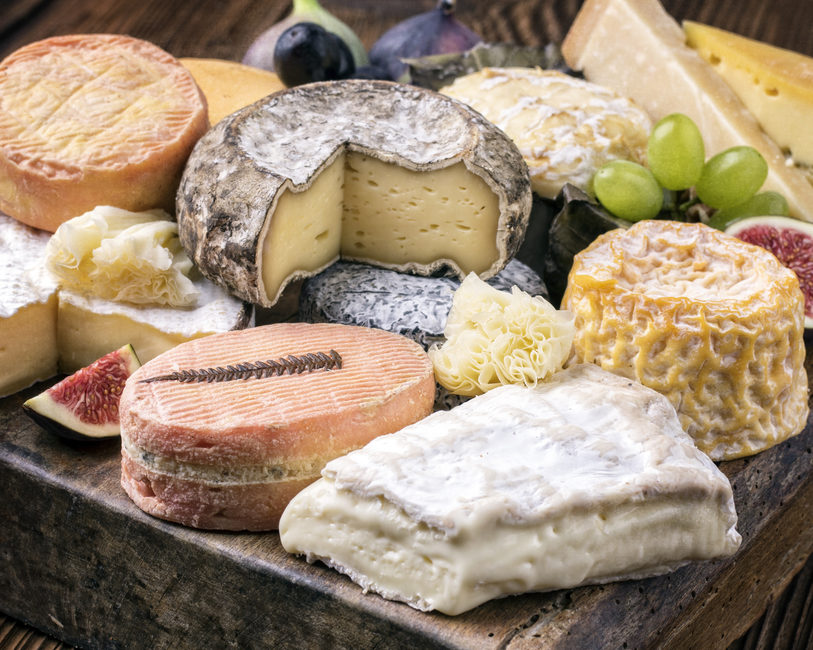 Le PLATEAU DE FROMAGE présente un assortiment de fromages artisanaux - à pâte molle, à pâte dure et à pâte vieillie - disposés sur une planche en bois avec des figues et des raisins, certains tranchés pour montrer les intérieurs crémeux et texturés.
