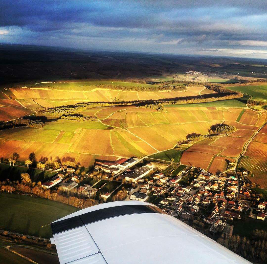 Vue aérienne de champs en patchwork et d'un village sous des nuages dramatiques, illuminés par la lumière du soleil, avec l'aile d'un avion au premier plan - parfait pour déguster le CHAMPAGNE ANDRÉ DILIGENT (Brut Blanc 75cl).