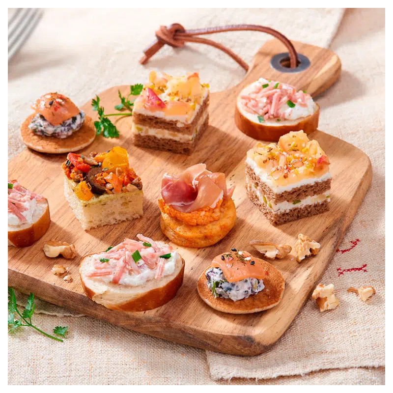 Le PETIT COCKTAIL FESTIF (13 pièces uniquement salées) présente une planche de service en bois avec des bouchées salées assorties, y compris du pain garni de viande, de fromage, d'herbes et de légumes sur un fond de tissu clair.