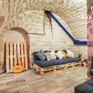 LE PETIT LOFT FACTORY (75019) propose un salon confortable avec des murs en pierre, un plafond voûté, des canapés et des coussins en palette, une palette en bois avec une guitare, des tuyaux bleus apparents et le logo de Magnolia Event dans le coin supérieur droit.