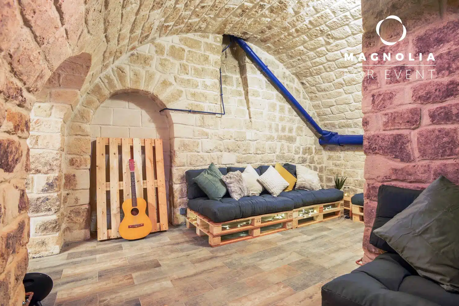 LE PETIT LOFT FACTORY (75019) propose un salon confortable avec des murs en pierre, un plafond voûté, des canapés et des coussins en palette, une palette en bois avec une guitare, des tuyaux bleus apparents et le logo de Magnolia Event dans le coin supérieur droit.