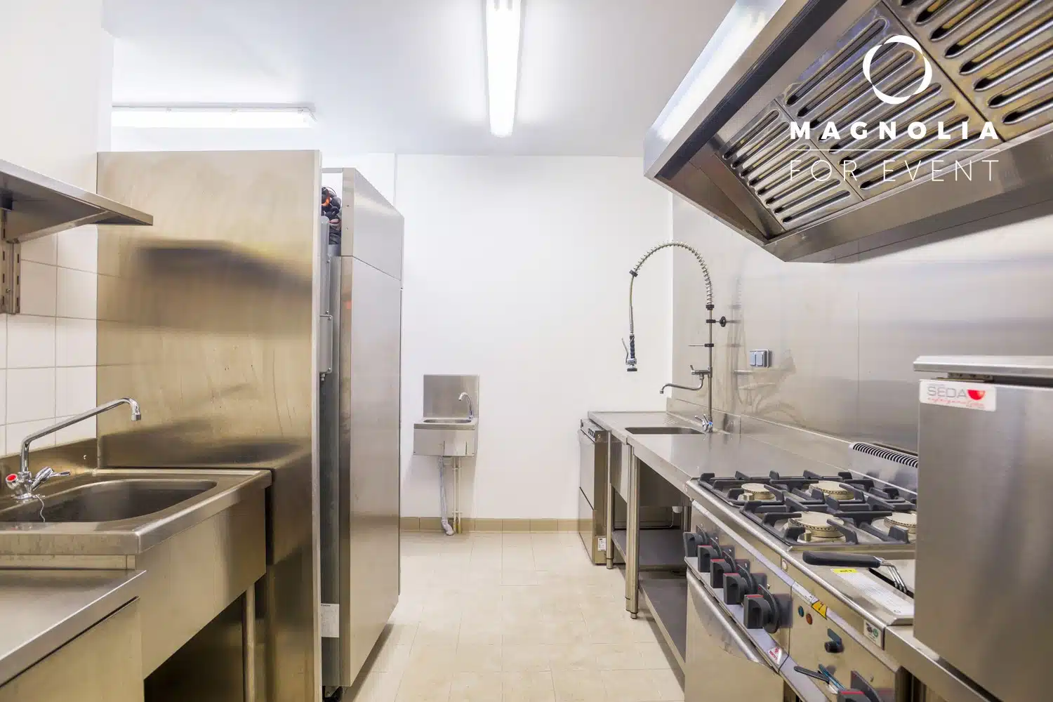 LE PETIT LOFT FACTORY (75019) dispose d'une cuisine moderne et élégante avec des appareils en acier inoxydable - cuisinière, évier, réfrigérateur et hotte de ventilation - brillamment éclairée par des plafonniers. Le logo de Magnolia Event est affiché dans le coin supérieur droit.