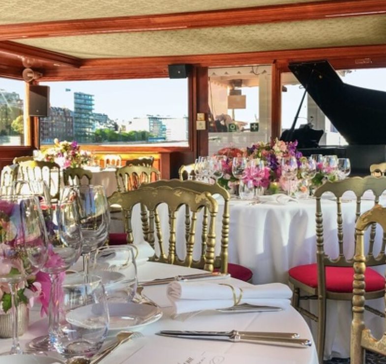 LE PETIT MISSISSIPI (75015) dispose d'une élégante salle à manger avec des tables rondes nappées de blanc, des chaises dorées avec des coussins rouges, des verres en cristal, des centres de table fleuris et un piano à queue noir près de la fenêtre qui donne sur les bâtiments de la ville.