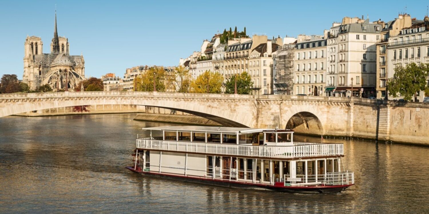 Le PETIT MISSISSIPI (75015), un bateau fluvial blanc, navigue sur la Seine à Paris avec les bâtiments historiques et la cathédrale Notre-Dame en arrière-plan sous un ciel bleu clair.