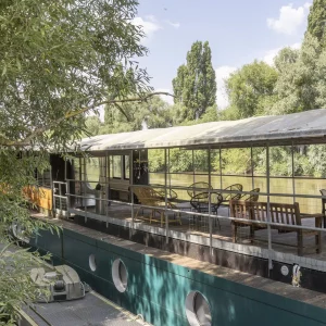 LA PÉNICHE DE LOUVECIENNES (78350) se trouve au bord d'une rivière calme, sa terrasse couverte est équipée de bancs et de chaises en bois, entourée d'arbres verdoyants sous un ciel bleu.