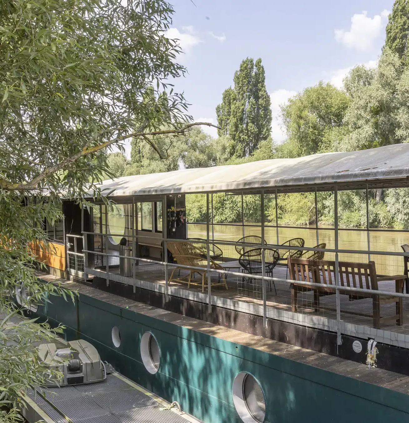 LA PÉNICHE DE LOUVECIENNES (78350) se trouve au bord d'une rivière calme, sa terrasse couverte est équipée de bancs et de chaises en bois, entourée d'arbres verdoyants sous un ciel bleu.