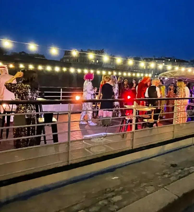 Un groupe en costumes et perruques colorés se réunit de nuit sur le pont de LA PÉNICHE NOTRE DAME DE PARIS (75005), éclairé par des guirlandes lumineuses, avec les bâtiments de la ville faiblement visibles en arrière-plan.