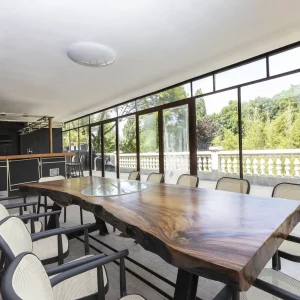 LE LOFT DE LA SEINE (78430) LOUVECIENNES offre un espace lumineux avec de grandes fenêtres, une table à manger en bois naturel avec des chaises, un coin cuisine moderne et des vues sur la verdure à l'extérieur.