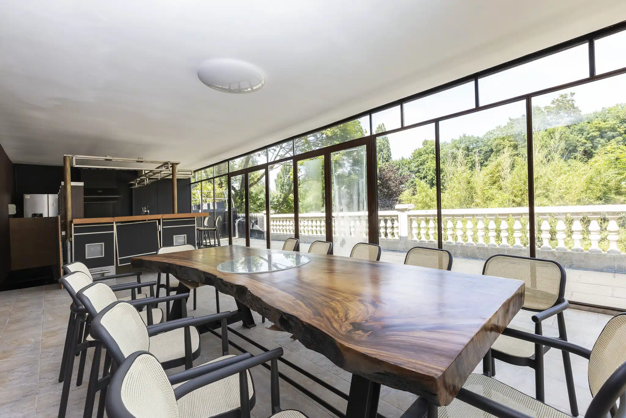 LE LOFT DE LA SEINE (78430) LOUVECIENNES offre un espace lumineux avec de grandes fenêtres, une table à manger en bois naturel avec des chaises, un coin cuisine moderne et des vues sur la verdure à l'extérieur.