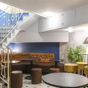 LE COSY CHIC présente un espace intérieur moderne avec des tables rondes, des tabourets bruns, une banquette brune touffetée près d'un mur bleu, un escalier avec une rampe lumineuse, des plantes en pot et des lampadaires contemporains.
