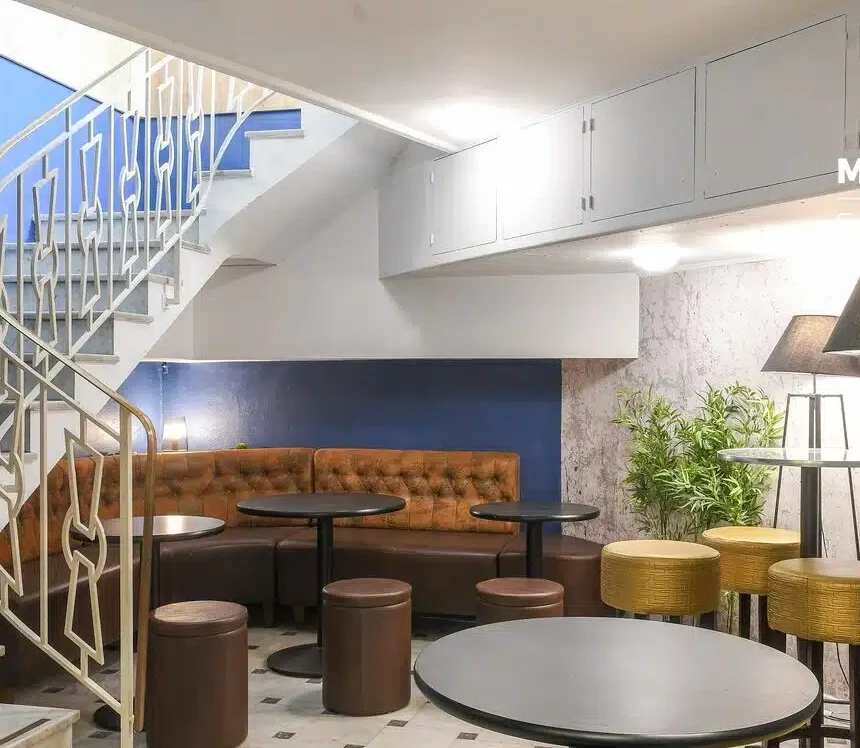 LE COSY CHIC présente un espace intérieur moderne avec des tables rondes, des tabourets bruns, une banquette brune touffetée près d'un mur bleu, un escalier avec une rampe lumineuse, des plantes en pot et des lampadaires contemporains.
