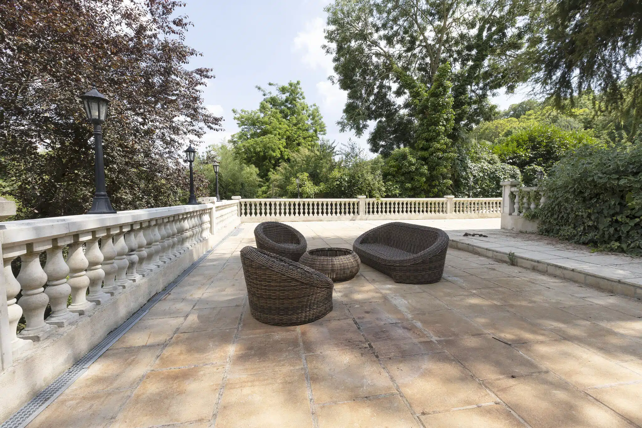 Le LOFT DE LA SEINE (78430) LOUVECIENNES dispose d'un patio extérieur avec des dalles en pierre beige, une balustrade blanche, des chaises en osier marron, une table ronde assortie, et est entouré de verdure sous un ciel partiellement nuageux.