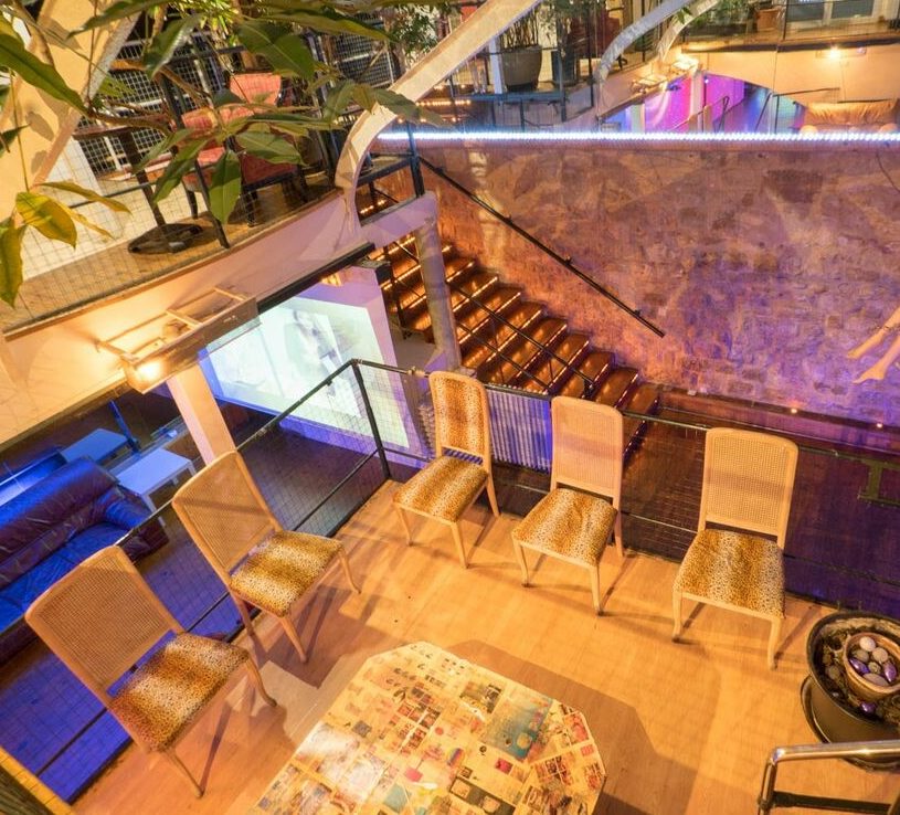 LE LOFT DE TITO (75011) propose un salon moderne et éclectique avec six chaises en osier près d'une table basse, un coin canapé confortable avec écran de projection, et un escalier éclairé qui surplombe l'espace.