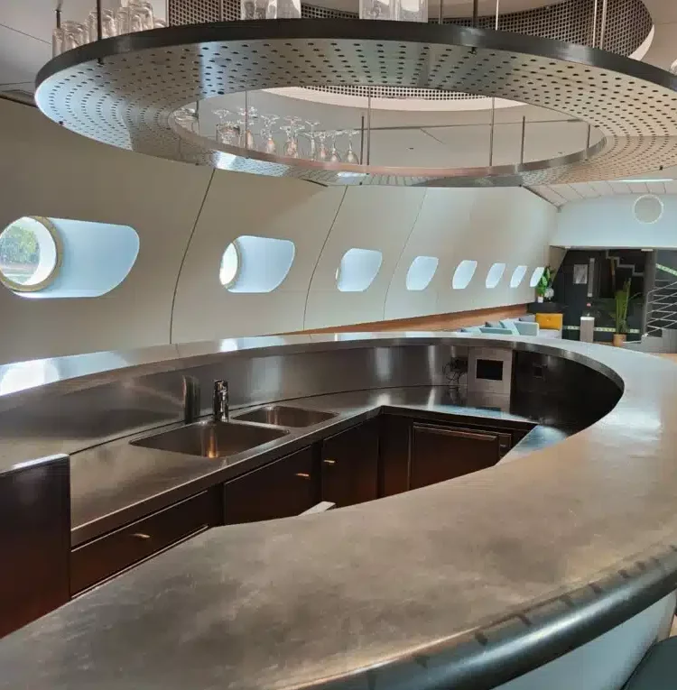 LE JAZZ HARMONY (75011) dispose d'un bar moderne incurvé avec un comptoir en acier inoxydable, un évier intégré, de grandes fenêtres ovales de style avion, un décor épuré et des étagères en verre au plafond.