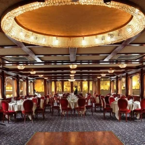 LE NASHVILLE TENNESSE (75015) présente une élégante salle à manger avec des chaises rouges, des tables rondes avec du linge blanc, des lustres en cristal, un plafond orné, de grandes fenêtres et une invitée en robe rouge qui passe devant les tables sur la droite.