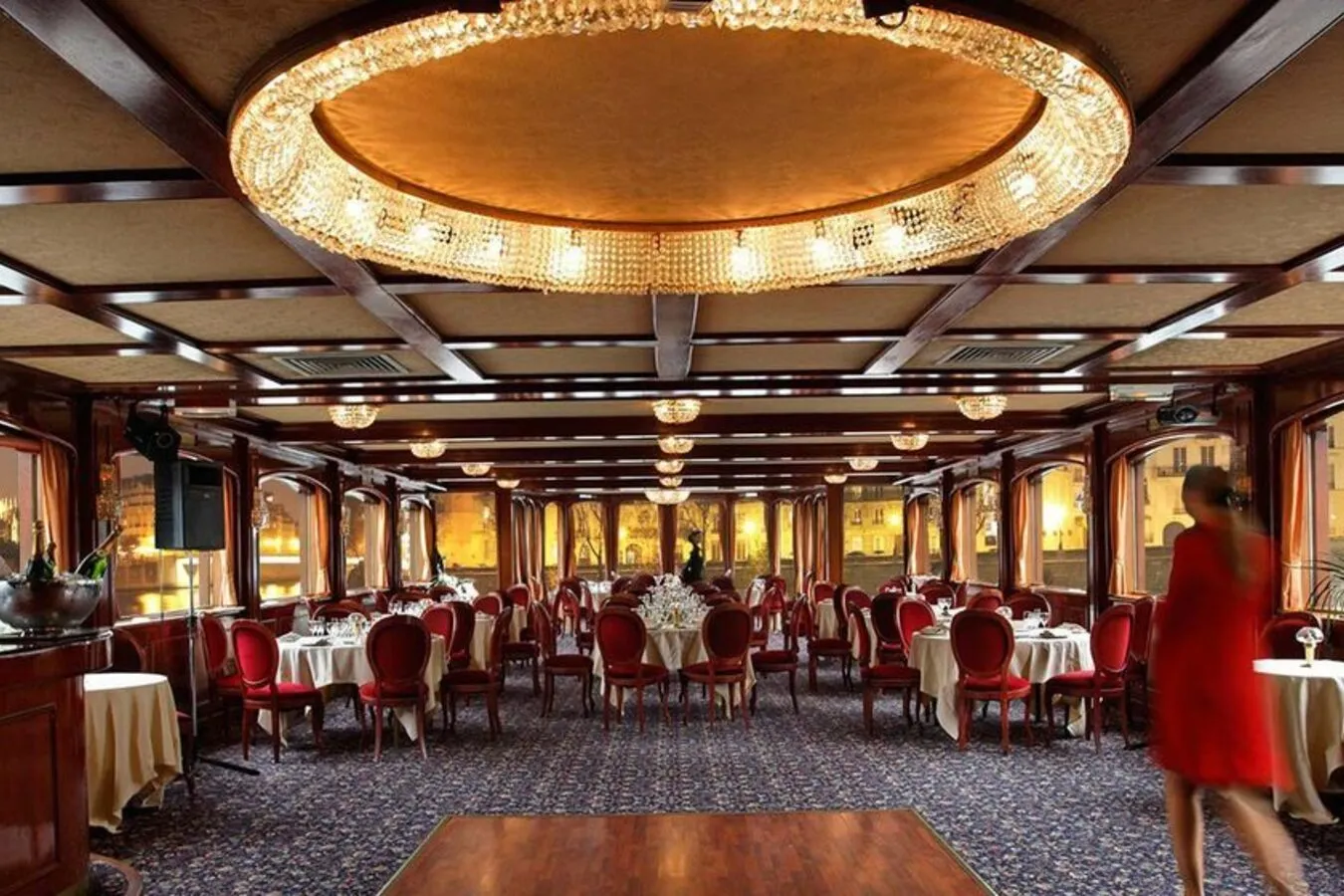 LE NASHVILLE TENNESSE (75015) présente une élégante salle à manger avec des chaises rouges, des tables rondes avec du linge blanc, des lustres en cristal, un plafond orné, de grandes fenêtres et une invitée en robe rouge qui passe devant les tables sur la droite.