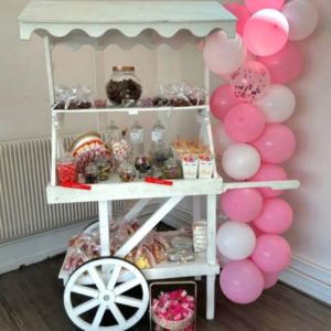 Le CANDY BAR SUR MESURE se compose d'un chariot en bois blanc rempli de bocaux et de sacs de bonbons colorés, agrémenté de colonnes de ballons roses et blancs pour une installation intérieure invitante.