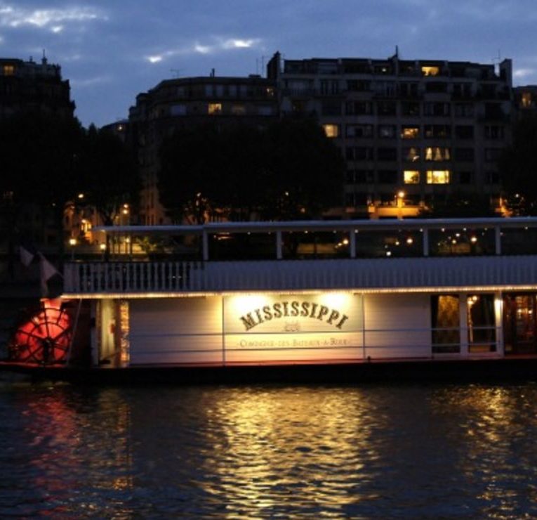 Le bateau fluvial LE PETIT MISSISSIPI (75015) brille de mille feux au crépuscule, amarré au bord de l'eau, avec des bâtiments urbains et des arbres derrière lui. Un ciel nuageux et sombre reflète les lumières de la ville sur le fleuve.