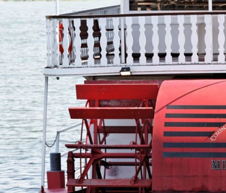 Un gros plan du LE PETIT MISSISSIPI (75015) met en valeur sa roue à aubes rouge, son design classique et ses détails mécaniques, sur fond de rambardes blanches et d'eau calme.