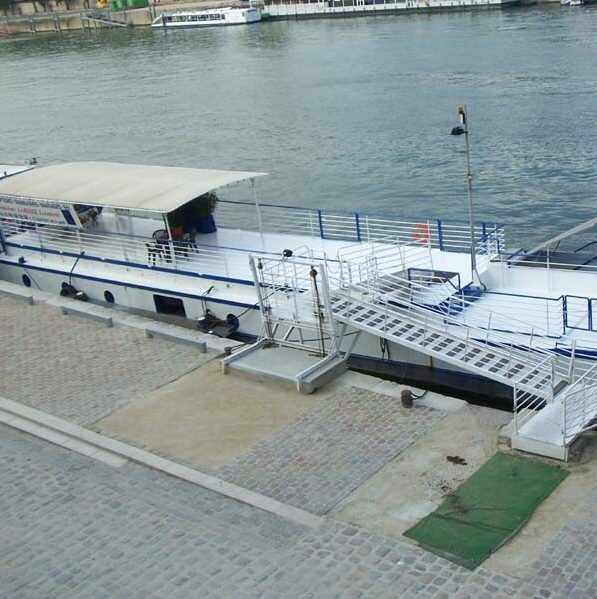 LA PÉNICHE ABYSSE (75013), un bateau blanc et bleu, est amarré le long d'une berge pavée avec une rampe d'accès à la rive ; on aperçoit les eaux calmes de la rivière et des bâtiments à l'arrière-plan.