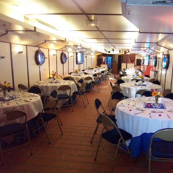 LA PÉNICHE ABYSSE (75013) dispose d'une salle de banquet avec des tables rondes drapées de blanc, équipées de verres et d'ustensiles. Des chaises noires entourent les tables, tandis que des fenêtres bordent les murs, créant ainsi un espace accueillant.