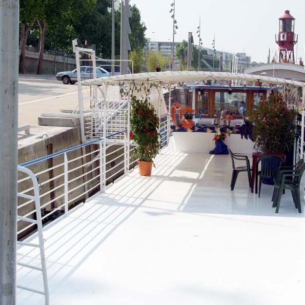 LA PÉNICHE ABYSSE (75013) présente un bateau à quai avec un pont blanc, des sièges couverts, des plantes en pot et des tables fleuries au bord de la rivière, avec un phare rouge visible à l'arrière-plan.