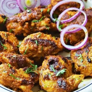 Achari Murg Tikka (300 g) : Morceaux de poulet épicés, dorés et grillés, à la texture croustillante et carbonisée, garnis d'herbes hachées et servis avec des oignons rouges émincés.